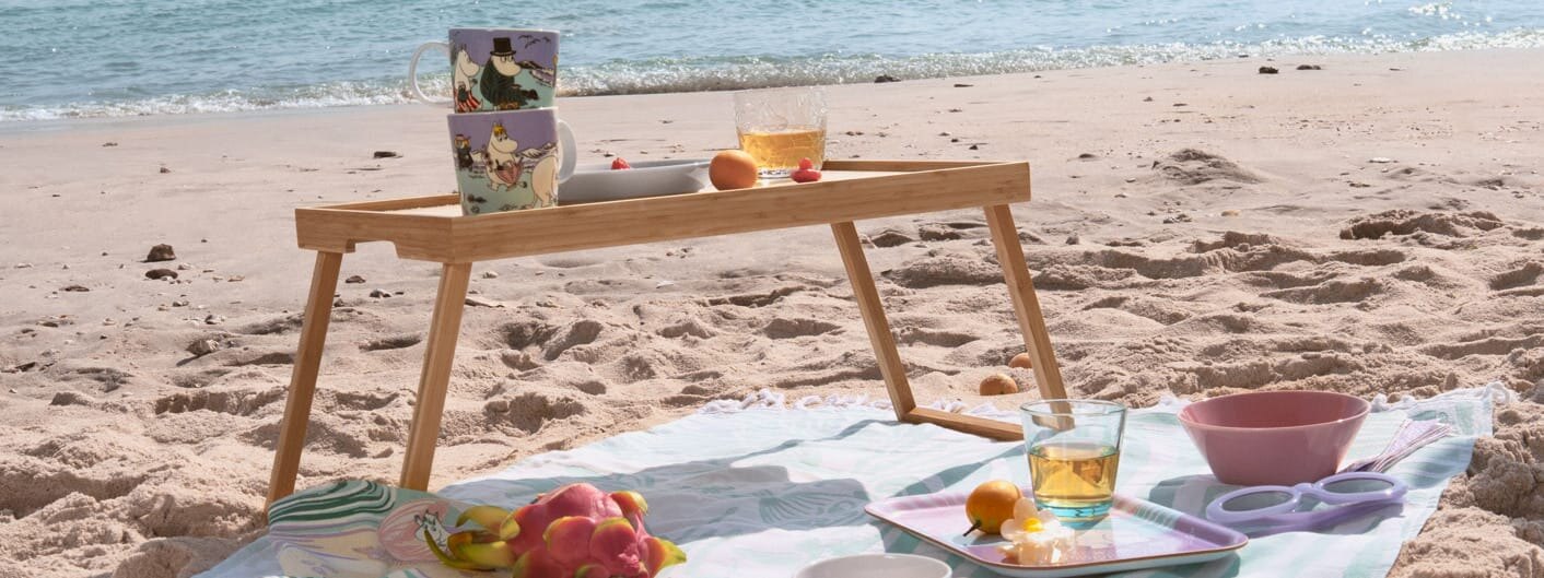 Moomin Arabia stranddag kopper og asjetter utstilt på picnic-teppe og brett på en sandstrand.