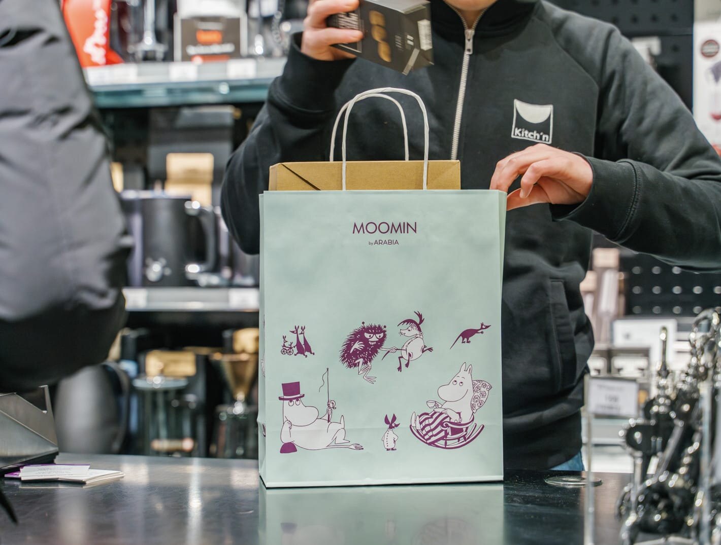 En butikkansatt hos Kitch'n står ved disken og legger varer oppi en Mummi-pose til en kunde.